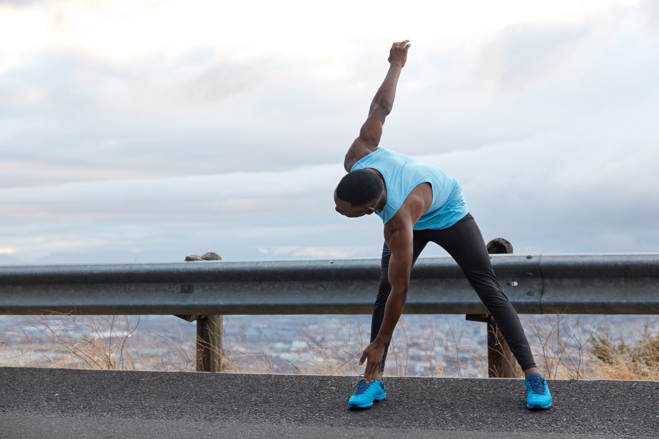 Horizontal shot of dark skinned man leans to feet, does stretching exercises, has muscular body shape, stands on asphalt over light blue sky with copy space for your advertising content, text